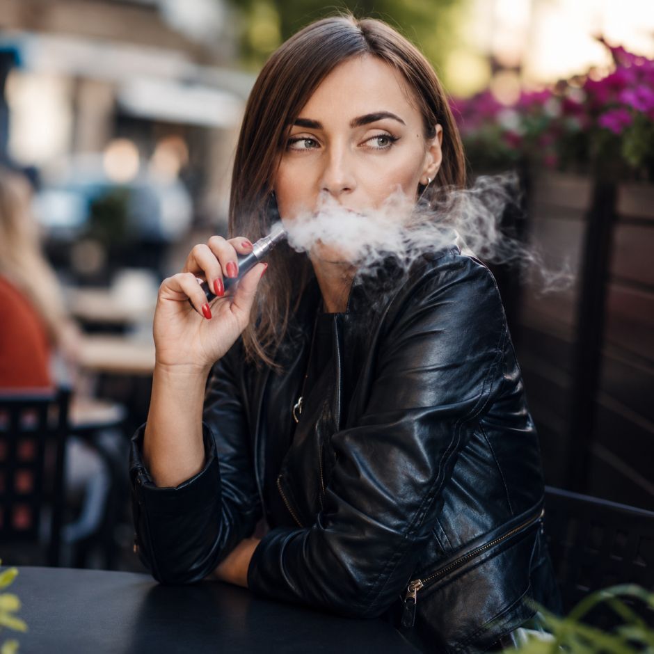 Adult using a vape device while seated outdoors, exhaling a light vapour cloud.